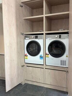 A laundry room with two front-loading washing machines, wooden cabinets, and a tiled floor. - Hidden Heritage Loft – Architect-Designed Northbridge Stay (Northbridge)