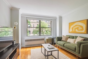 A living room with a green sofa, a coffee table, a television, and a large window with a view of buildings and trees. - Blueground | Central Park West, gym, nr Central Park (New York)