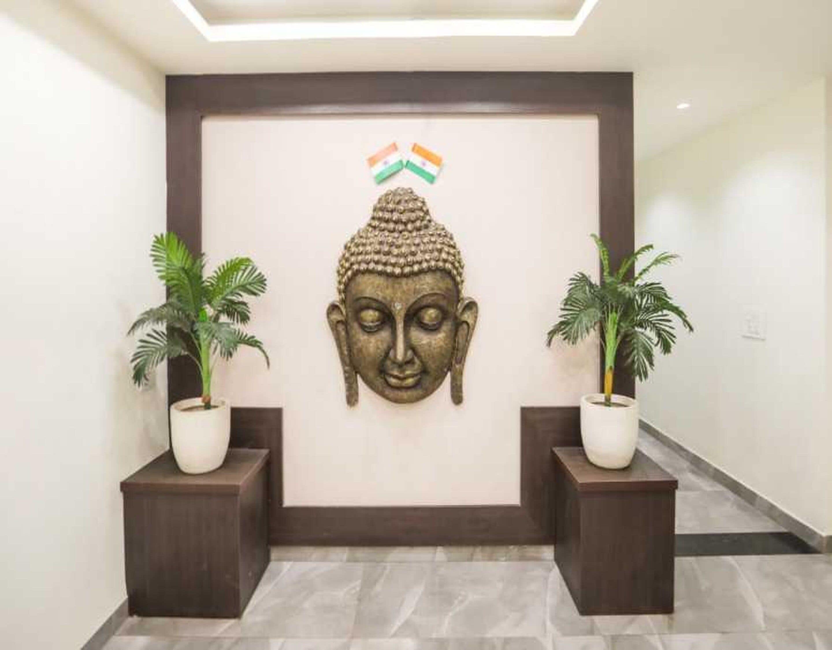Una escultura de cabeza de Buda montada en la pared, con un lazo tricolor encima, flanqueada por dos macetas con plantas sobre pedestales oscuros en un pasillo.