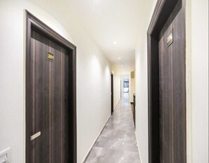 A hallway with dark wooden doors, each marked with a number. The floor is tiled, and the walls are painted white. - Deluxe Hotel Krishna (Faridabad)