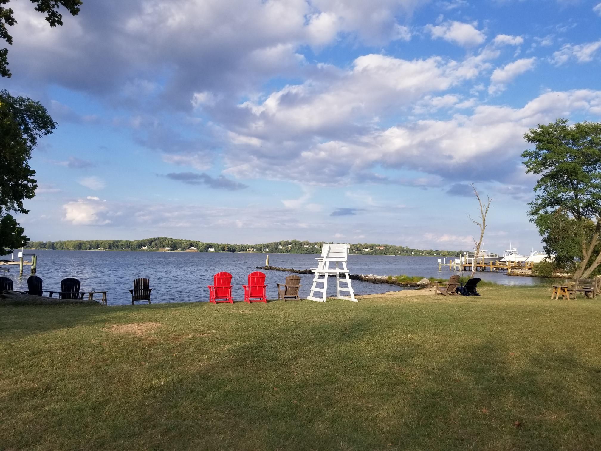 Vista al lago con zona de césped, varias sillas Adirondack y un puesto de salvavidas blanco.