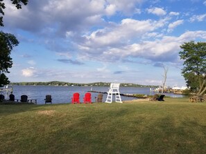A lakeside view with a grassy area, several Adirondack chairs, and a white lifeguard stand.