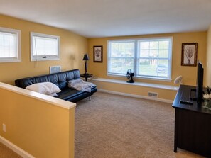 A living room with a black leather sofa, a small table with a lamp, a large window with blinds, and a television on a stand. - Waterfront Estate Renting 4BR Guest Cottage w/waterviews of Magothy River (Arnold)