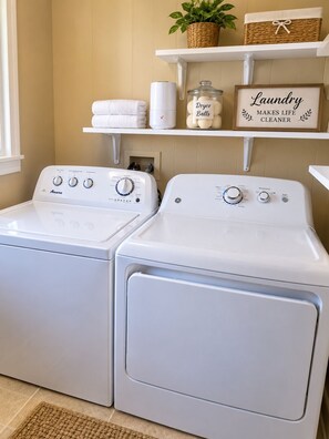 A laundry area with two white front-loading washing machines, a shelf with towels and a sign saying 'Laundry Makes Life Cleaner', and a basket on the wall.