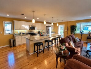 A modern kitchen with a central island, bar stools, and a dining area with a brown sofa and a small table. - Waterfront Estate Renting 4BR Guest Cottage w/waterviews of Magothy River (Arnold)