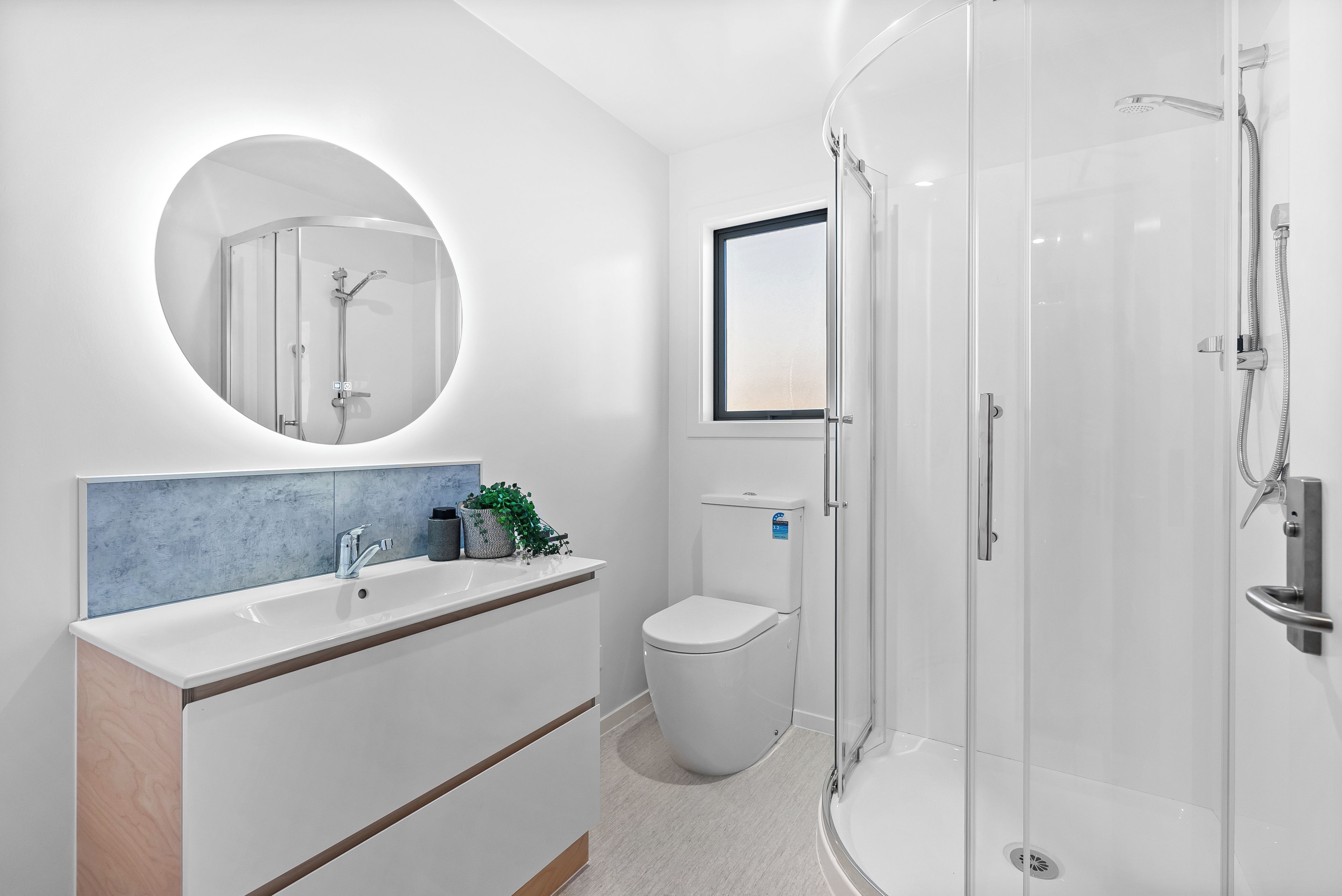 A modern bathroom with a round mirror, a white sink, a toilet, and a glass-enclosed shower area.
