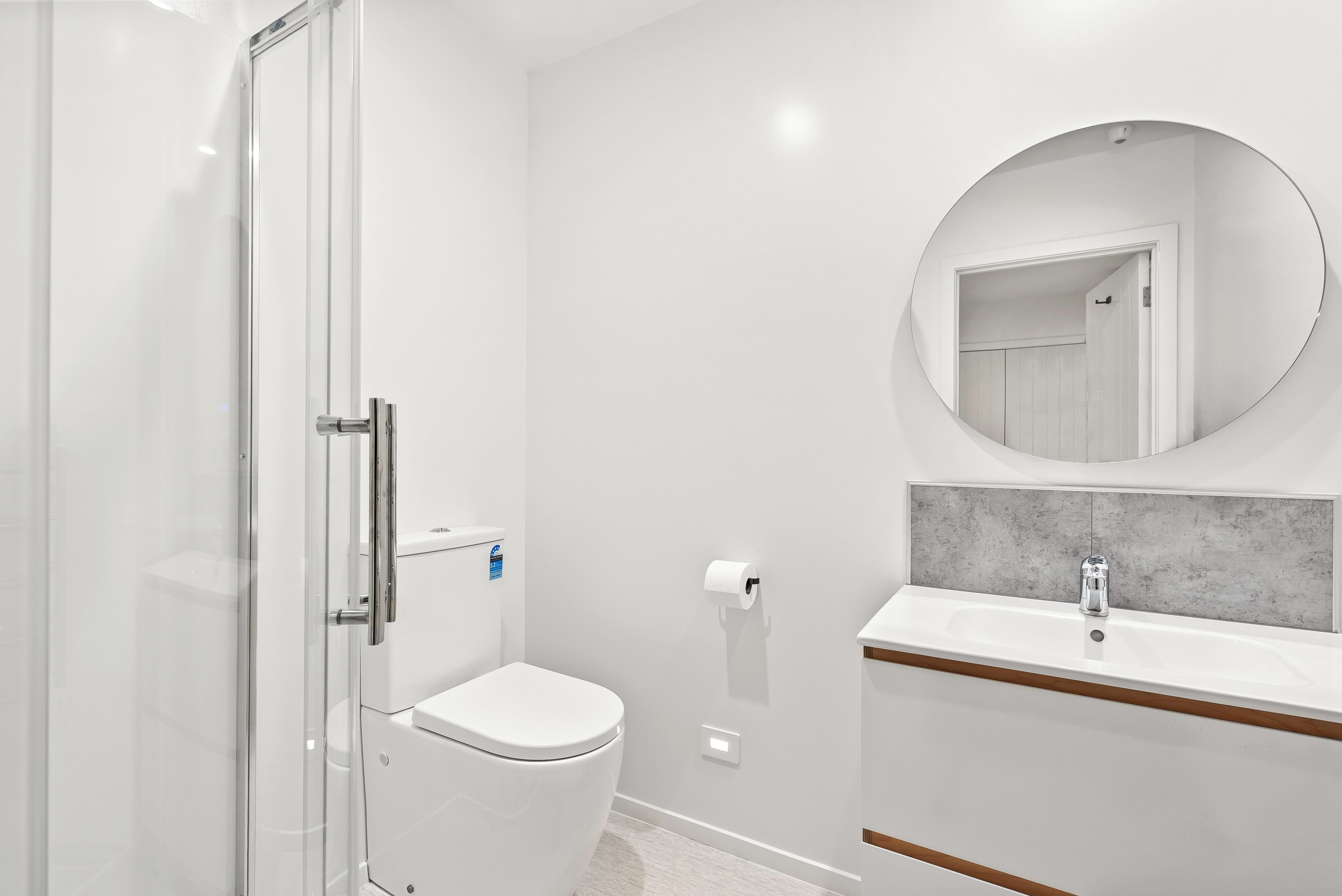 A modern bathroom with a glass shower enclosure, a toilet, a sink with a marble countertop, and a round mirror.