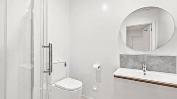 A modern bathroom with a glass shower enclosure, a toilet, a sink with a marble countertop, and a round mirror.