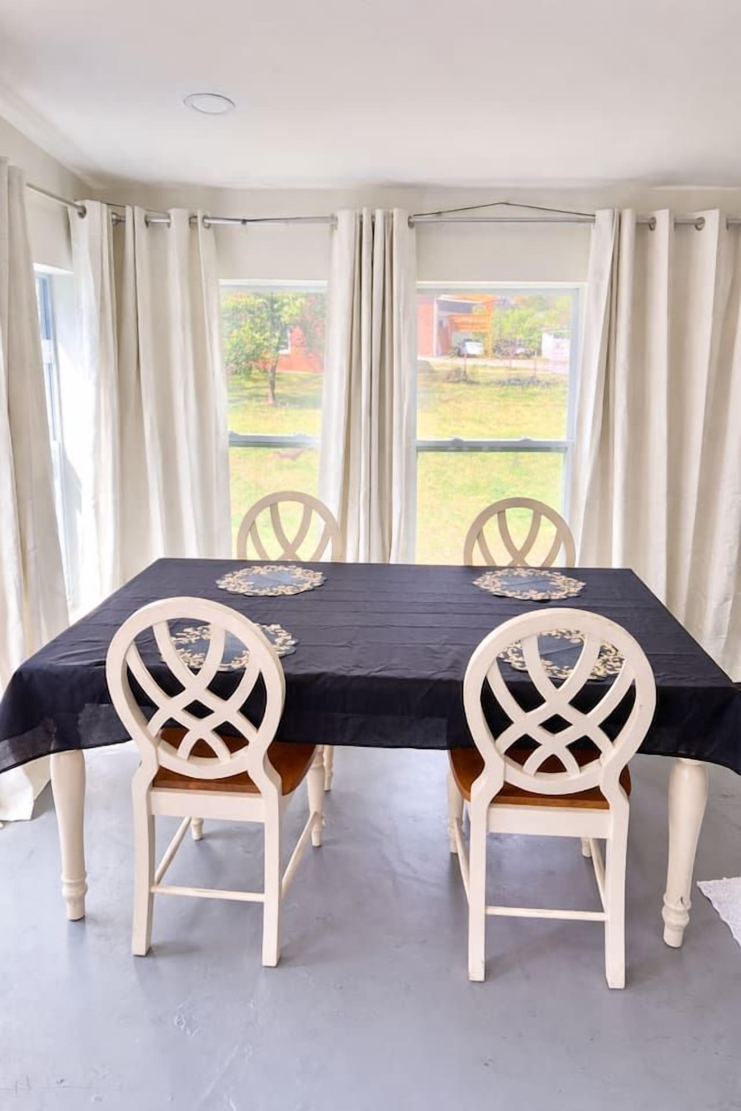 A dining table with a black tablecloth and white chairs, positioned in a room with large windows and curtains.