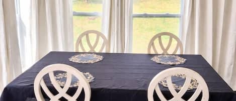 A dining table with a black tablecloth and white chairs, positioned in a room with large windows and curtains.