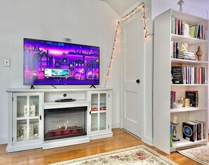 A living room with a flat-screen TV on a white entertainment unit, a fireplace, a bookshelf, and a door with a string of lights. - Romantic Fairytale |10 min to Downtown (East Point)