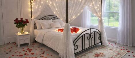 A four-poster canopy bed with rose petals on the floor, a bedside table with a vase of red roses, and a window with curtains.