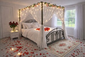 A four-poster canopy bed with rose petals on the floor, a bedside table with a vase of red roses, and a window with curtains.