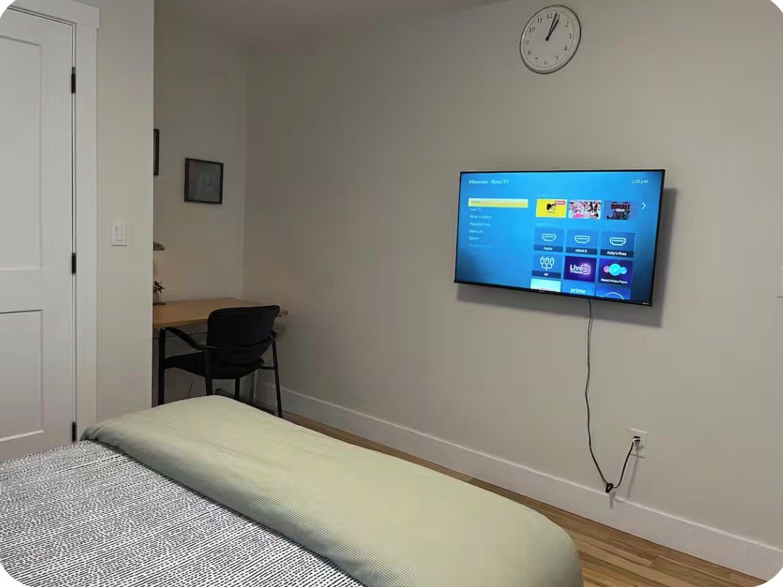 Ein Zimmer mit einem Bett, einem Schreibtisch, einem Stuhl und einem an der Wand befestigten Fernseher, der einen Homescreen anzeigt.