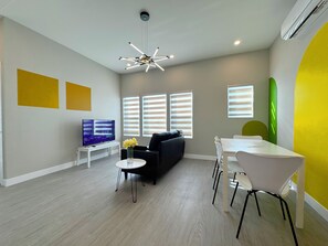 A modern living room with a flat-screen TV, a black sofa, a dining table with chairs, and a unique ceiling light fixture. - 5704- Cozy 2B1B Private Unit near FWY 580/24 & UCB (Oakland)