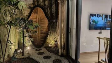 Un interior moderno con una puerta de madera, un pequeño jardín con piedras y plantas, y un televisor montado en la pared.