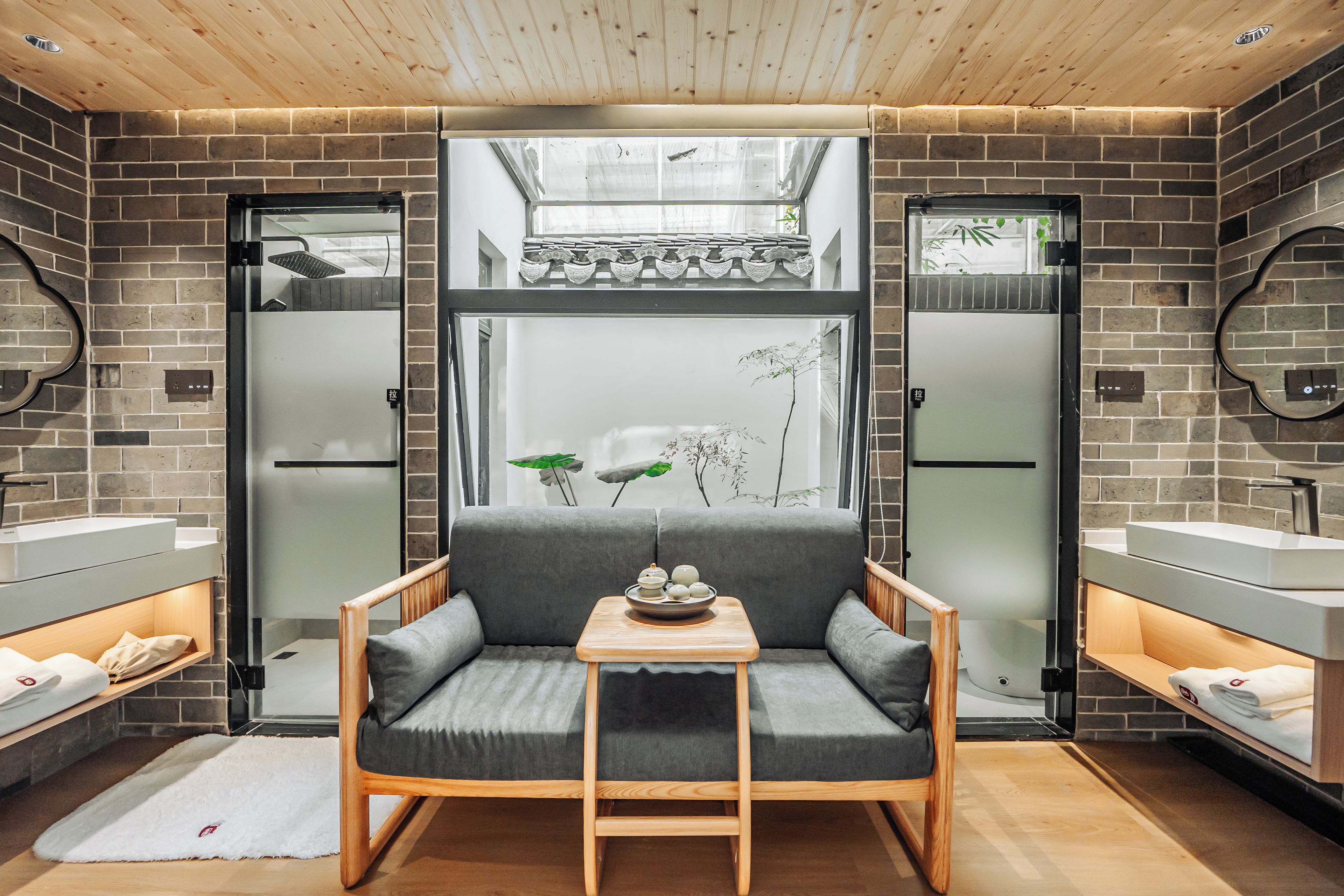 Un baño moderno con sofá gris, dos lavamanos y una ventana con vistas a la vegetación.