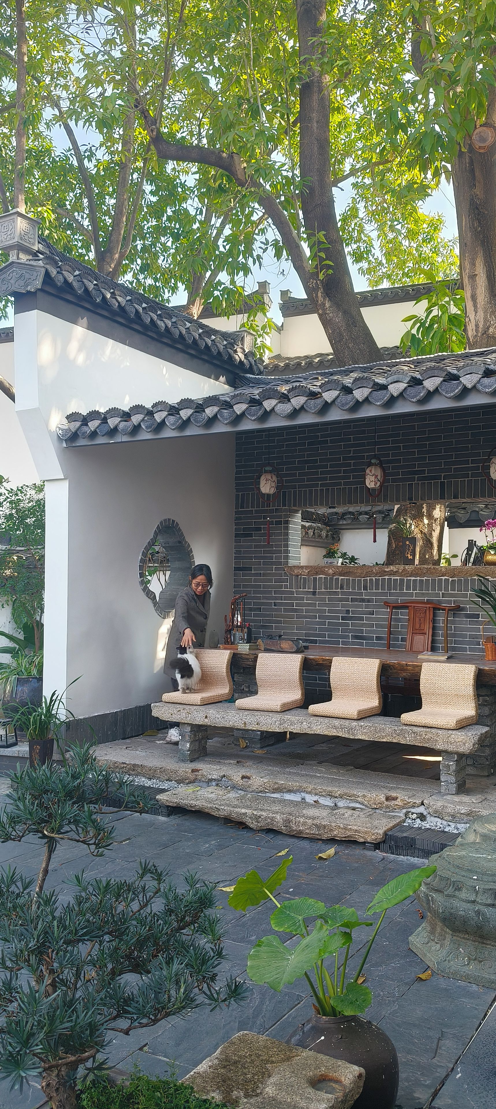 A traditional-style building with a tiled roof and brick walls, a stone bench, and potted plants.