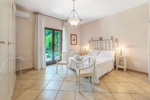 Une chambre à coucher comprenant un lit, deux tables de chevet, une chaise, une table, une fenêtre avec des rideaux et une vue sur la verdure à l’extérieur.
