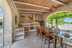 A stone-walled kitchen with a built-in oven, a dining area with wooden tables and chairs, and a view of a beach. - Masseria Saracino Pool And Garden, Martano, Italy (Martano)