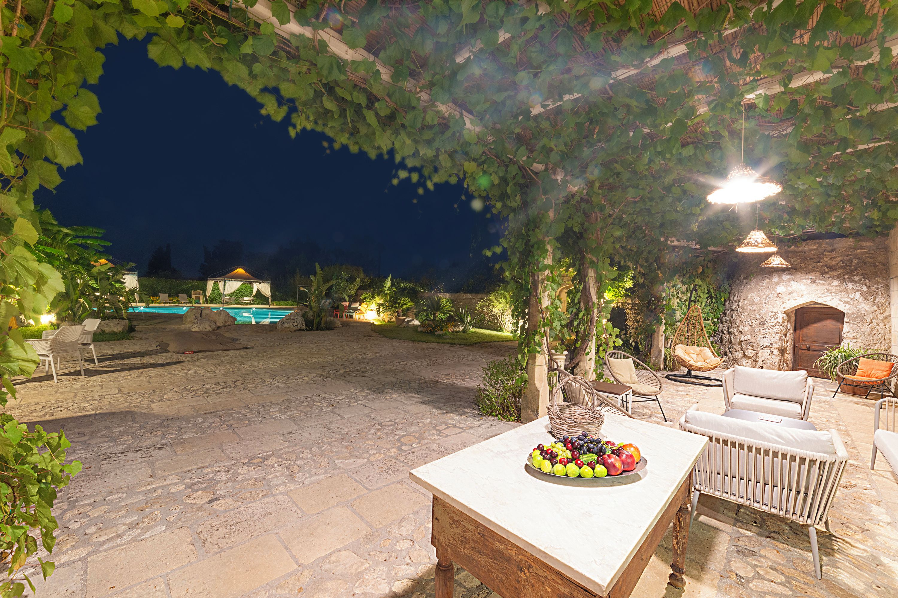 Un patio al aire libre con suelo de piedra, una mesa con fruta, muebles de mimbre y una piscina al fondo.