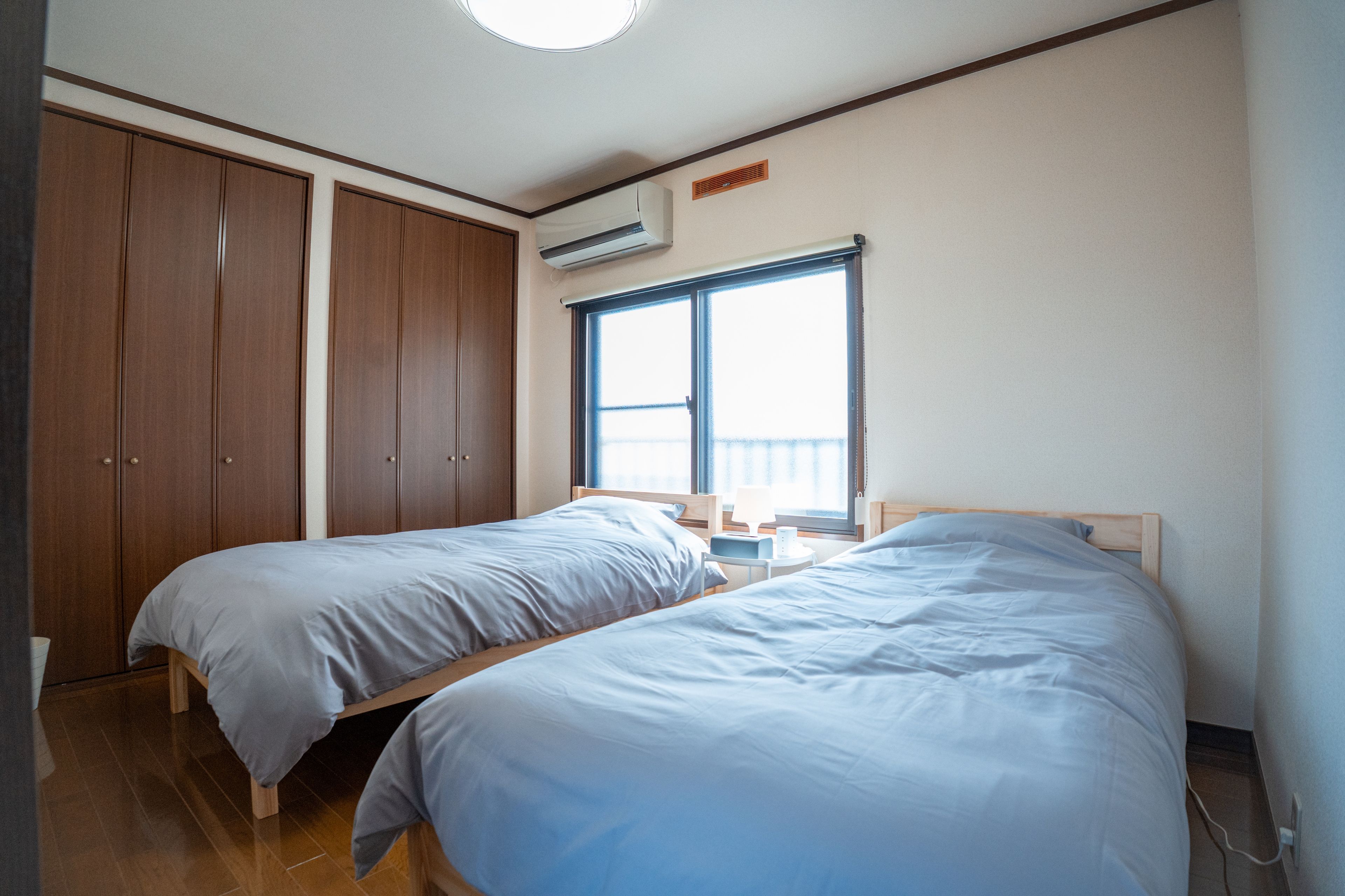 A bedroom with two single beds, a wardrobe, a window with blinds, and an air conditioning unit.