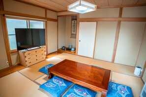 Ein traditioneller japanischer Raum mit Tatami-Bodenbelag, einem Holztisch, einem Fernseher und einer Schiebetür.