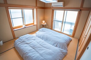 Ein Zimmer mit einem Bett, zwei Fenstern, einem kleinen Tisch mit einer Lampe und einer Klimaanlage.