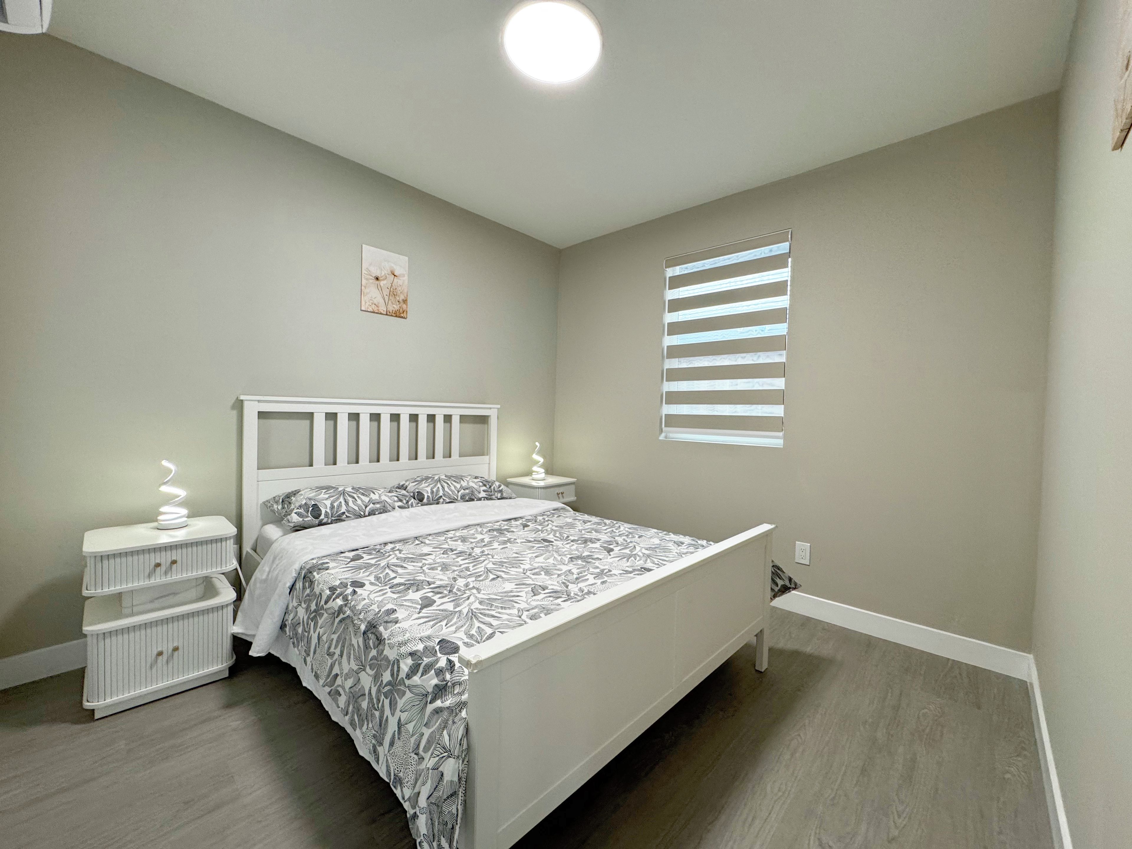 A bedroom with a bed, bedside tables, a wall-mounted light, a window with blinds, and a small picture on the wall.