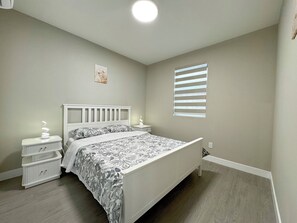 Ein Schlafzimmer mit einem Bett, Nachttischen, einer Wandleuchte, einem Fenster mit Jalousien und einem kleinen Bild an der Wand.