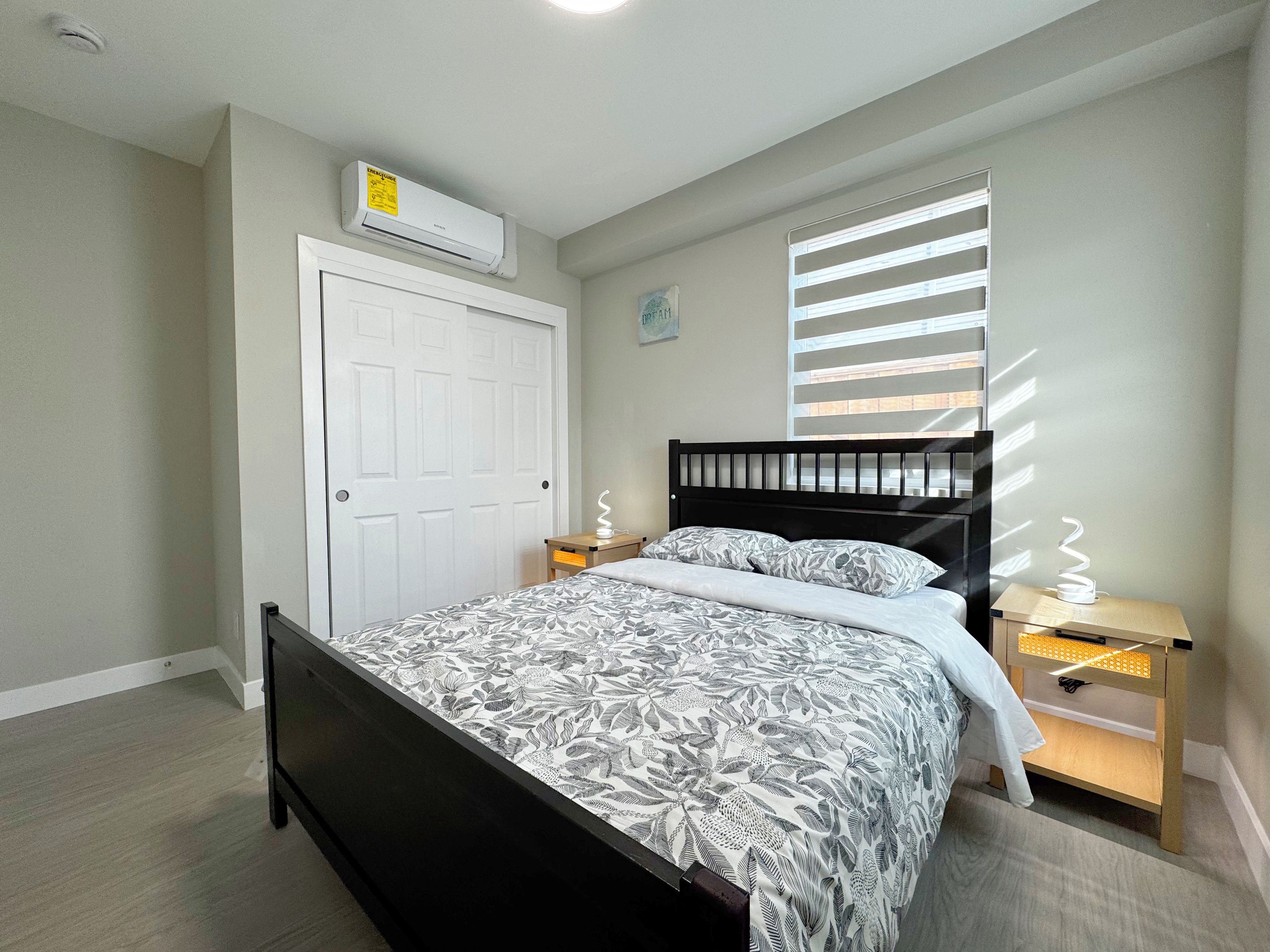 A bedroom with a bed, a white door, a window with blinds, a bedside table with a lamp, and an air conditioning unit.
