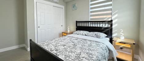 A bedroom with a bed, a white door, a window with blinds, a bedside table with a lamp, and an air conditioning unit.