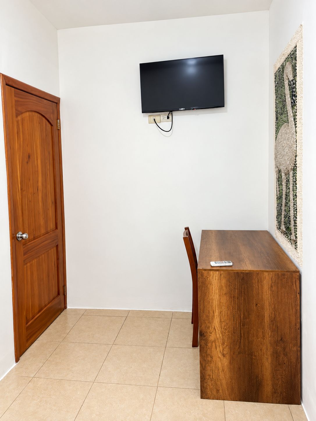 Quarto com porta de madeira, televisão de tela plana na parede, mesa de jantar de madeira com cadeiras e um objeto decorativo na parede.