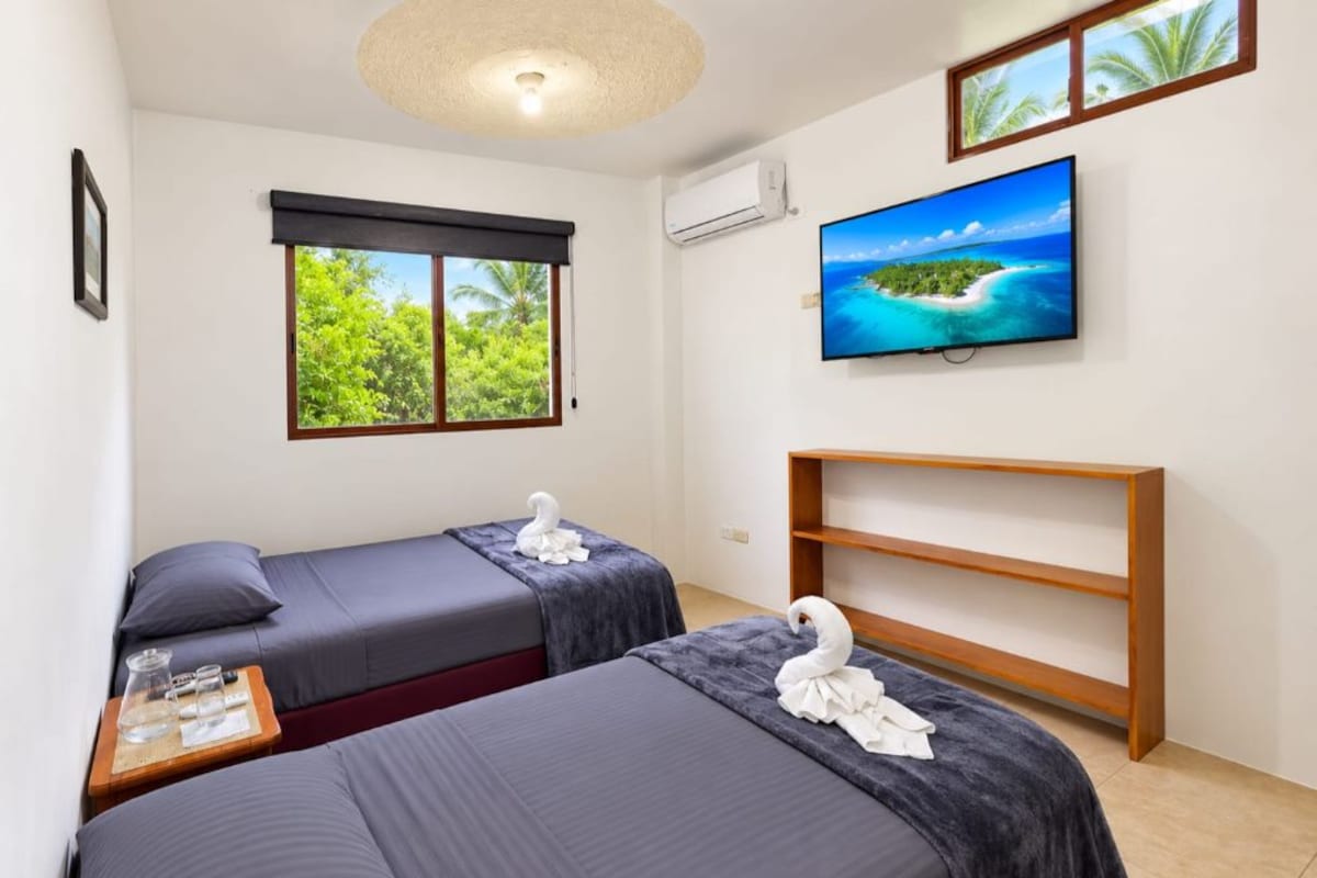 A hotel room with two beds, a TV, a window with a view of greenery, and a shelf.