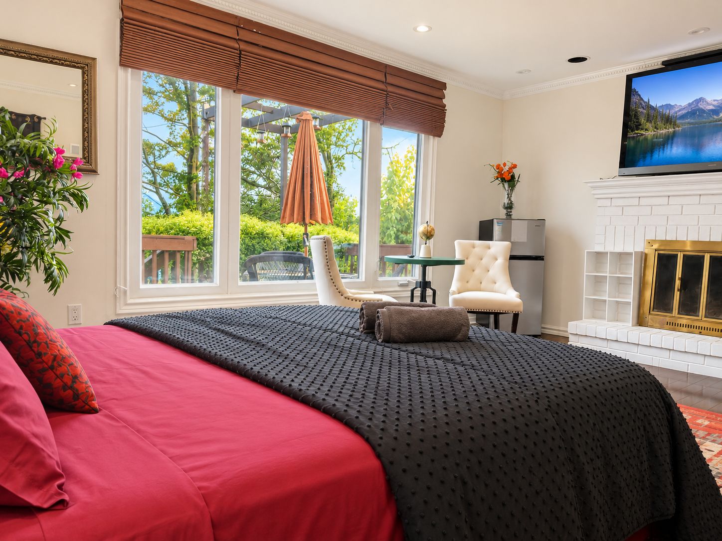 A bedroom with a large bed, a television, a fireplace, and a view of greenery outside.