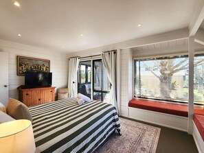 A bedroom with a bed, a television on a wooden cabinet, a window with curtains, and a view of the outside. - The Heart of Hahei, a modern and central home only steps away from shops / beach (Hahei)