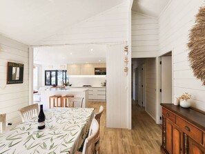 A modern kitchen with white cabinetry and a central island, a dining area with a table set for two, and a hallway leading to other rooms. - The Heart of Hahei, a modern and central home only steps away from shops / beach (Hahei)