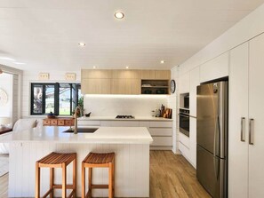 A modern kitchen with white cabinetry, a stainless steel refrigerator, and wooden stools. - The Heart of Hahei, a modern and central home only steps away from shops / beach (Hahei)