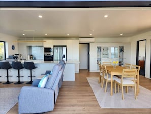 A modern kitchen with a central island, bar stools, a dining table with chairs, and a living area with a sofa and a coffee table. - Tirohanga a Hei - "The view of Hei" Adults only (Hahei)