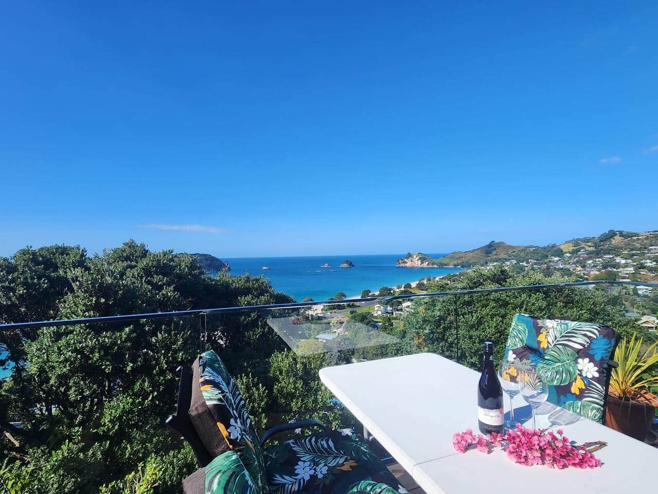 A balcony with a table, a bottle, and glasses, overlooking a coastal landscape with trees and buildings.