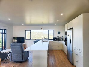A modern kitchen with a central island, white cabinetry, and a large window providing a view. - Tirohanga a Hei - "The view of Hei" Adults only (Hahei)
