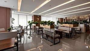 Un moderno vestíbulo de hotel con un diseño limpio y minimalista, que cuenta con un área de descanso central con mesas y sillas, una recepción y plantas decorativas.