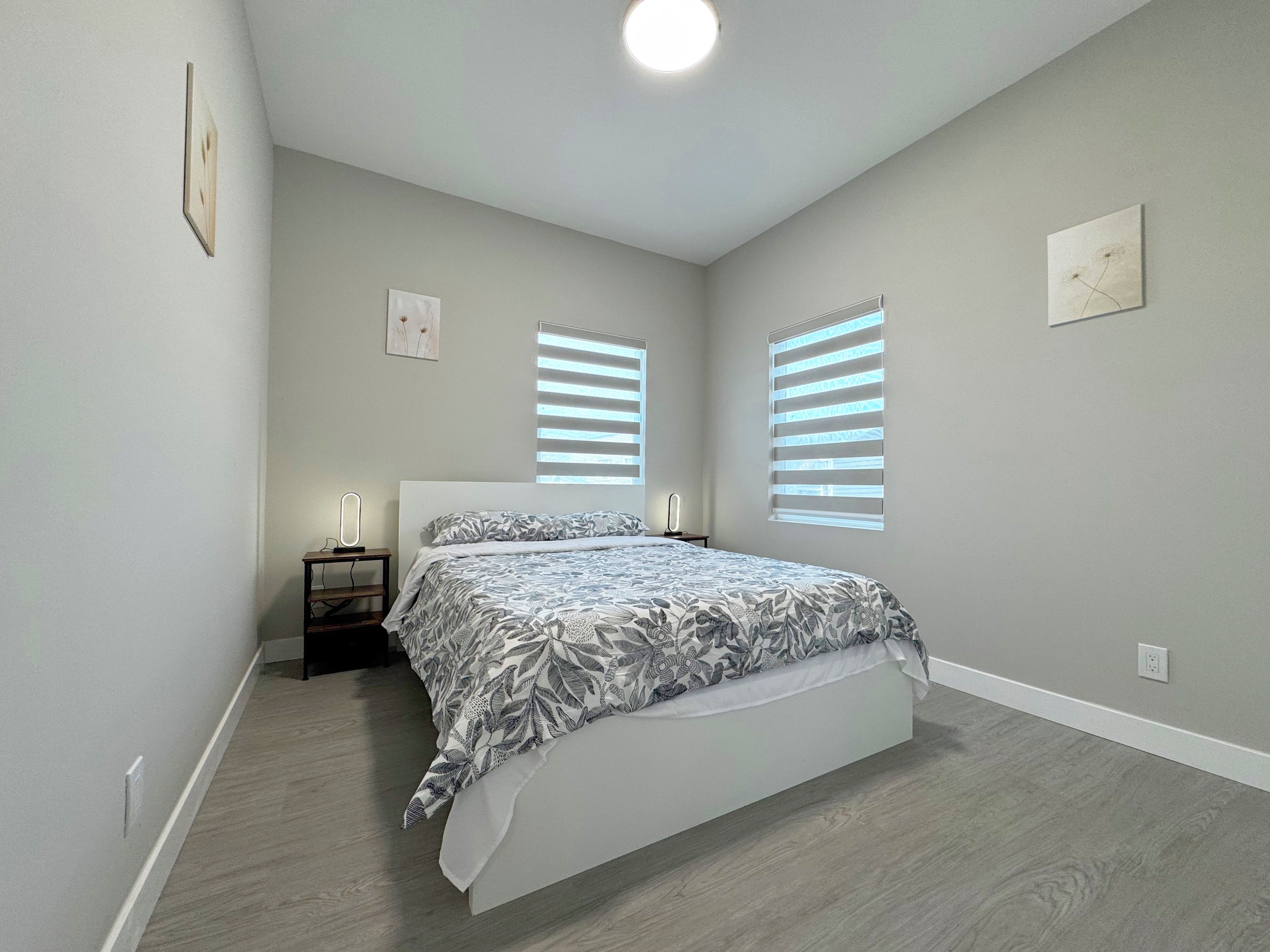 A bedroom with a bed, two bedside tables, and two windows with blinds.
