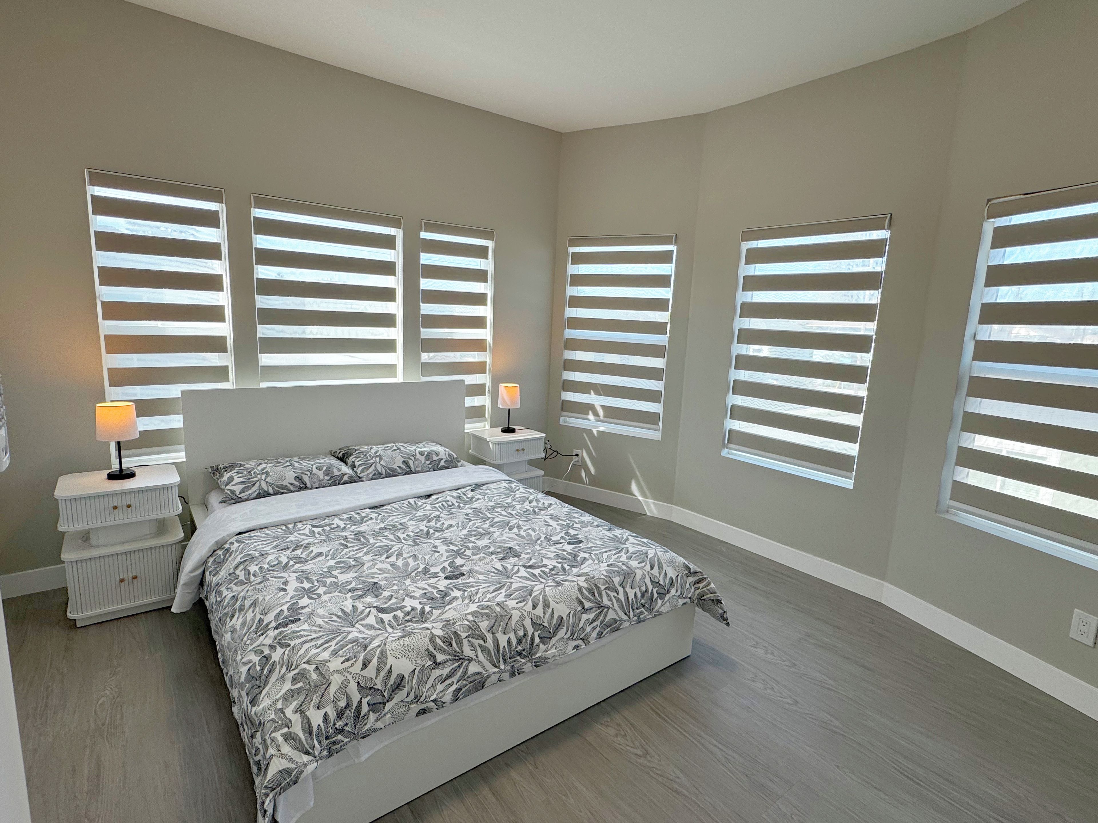A bedroom with a large bed, two bedside tables, and multiple windows with blinds.