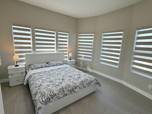 Ein Schlafzimmer mit einem großen Bett, zwei Nachttischen und mehreren Fenstern mit Jalousien.