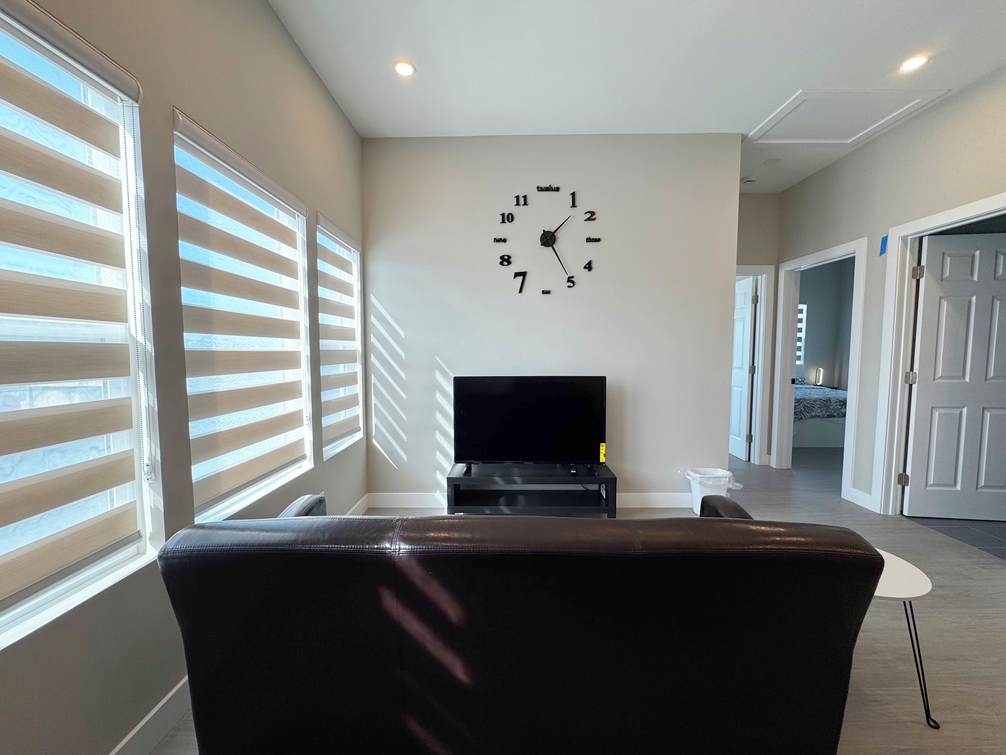 A living room with a black leather sofa, a flat-screen TV on a stand, and a wall clock.