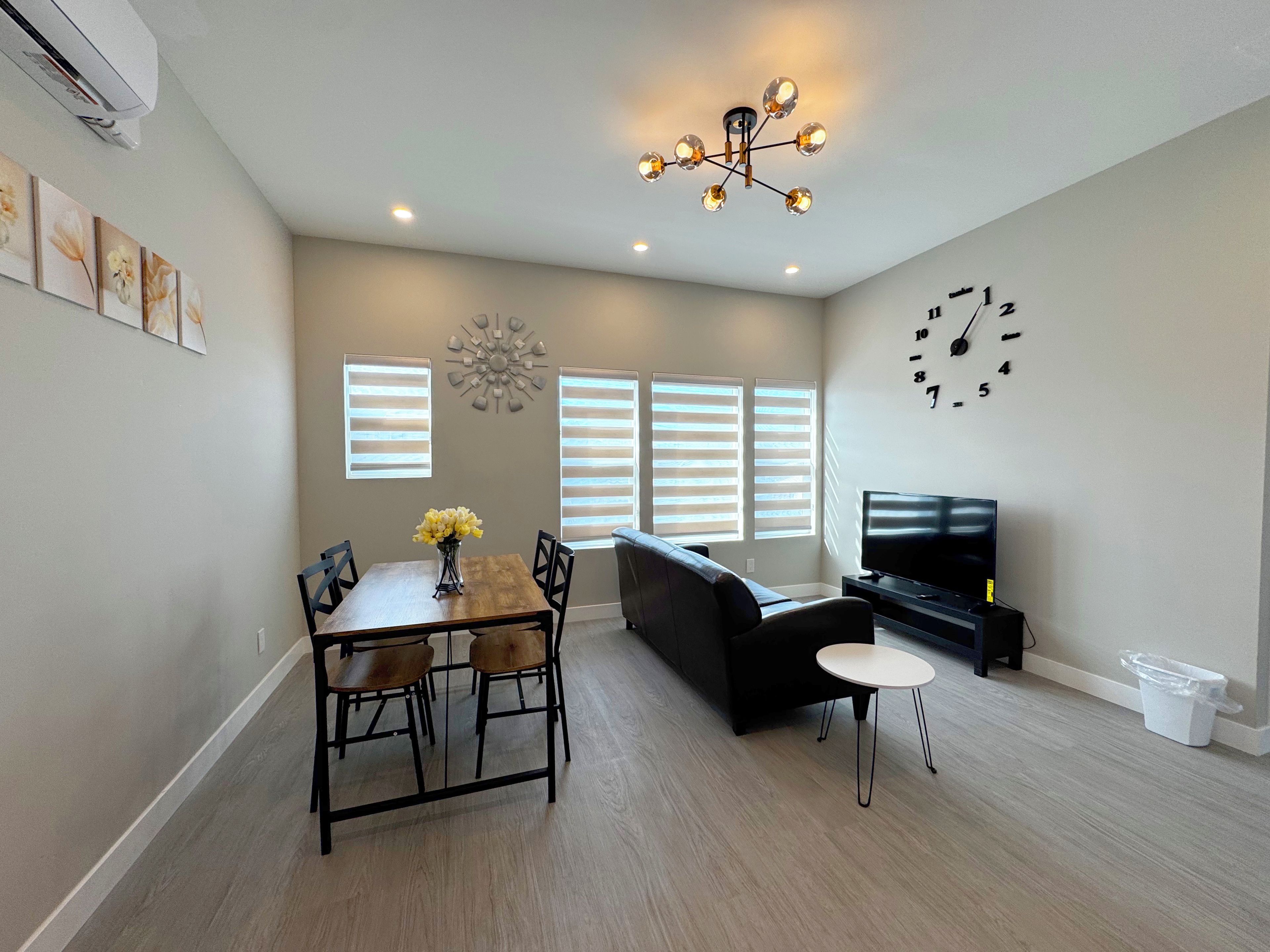 A modern living room with a dining area, a black sofa, a TV, and a clock on the wall.