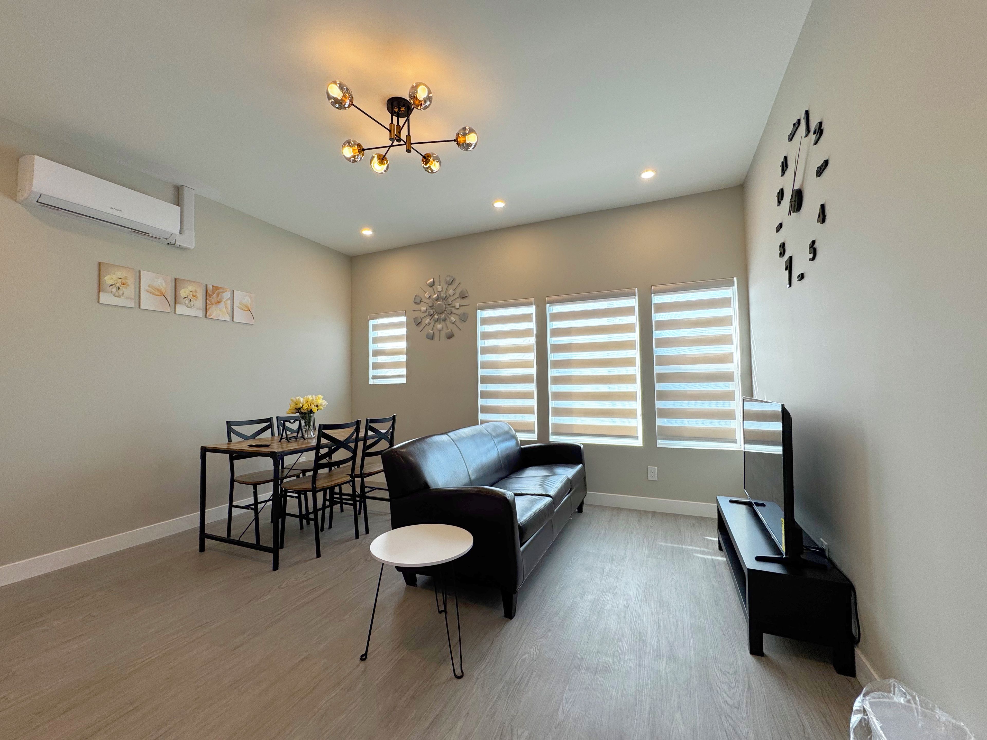 A living room with a sofa, a dining table with chairs, a small round table, a TV stand, and a ceiling light fixture.