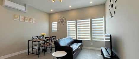 A living room with a sofa, a dining table with chairs, a small round table, a TV stand, and a ceiling light fixture.