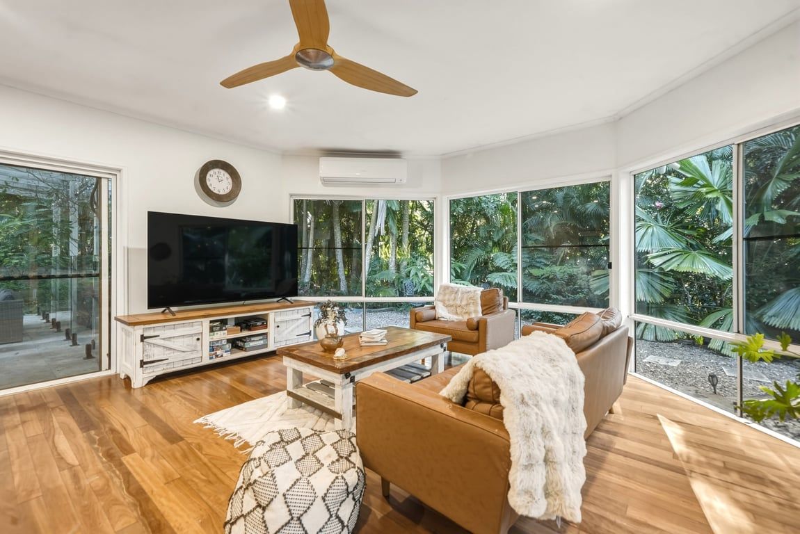 A modern living room with a flat-screen TV, wooden flooring, a ceiling fan, and large windows offering a view of greenery.